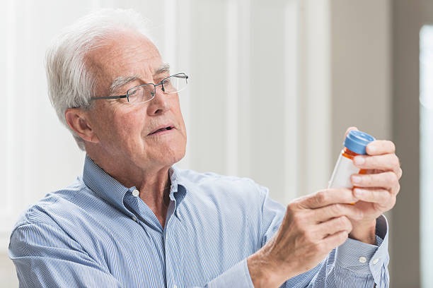 An older man in glasses squinting at the label on a pill bottle.